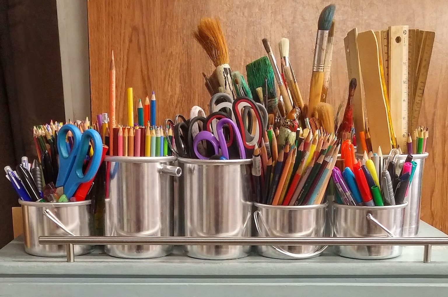 Various art supplies in metal containers on a wooden table including colored pencils, paint brushes, scissors, markers, and a ruler.